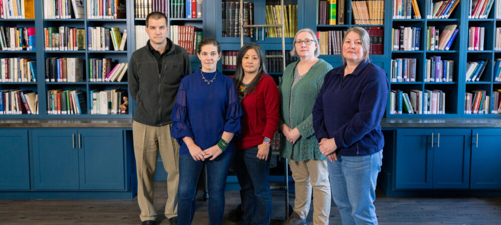 Five of the seven professors who make up the Gustavus Adolphus College Faculty Task Force stand with seriousness before a wall of books.