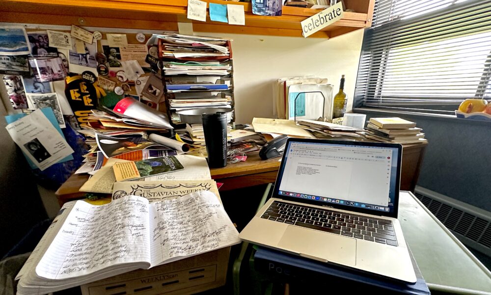 The messy desk of Gustavus Adolphus College professor and poet Phil Bryant ’73, who is writing poetry there with a notebook and a laptop propped up on a hardcover book titled "Poetry".